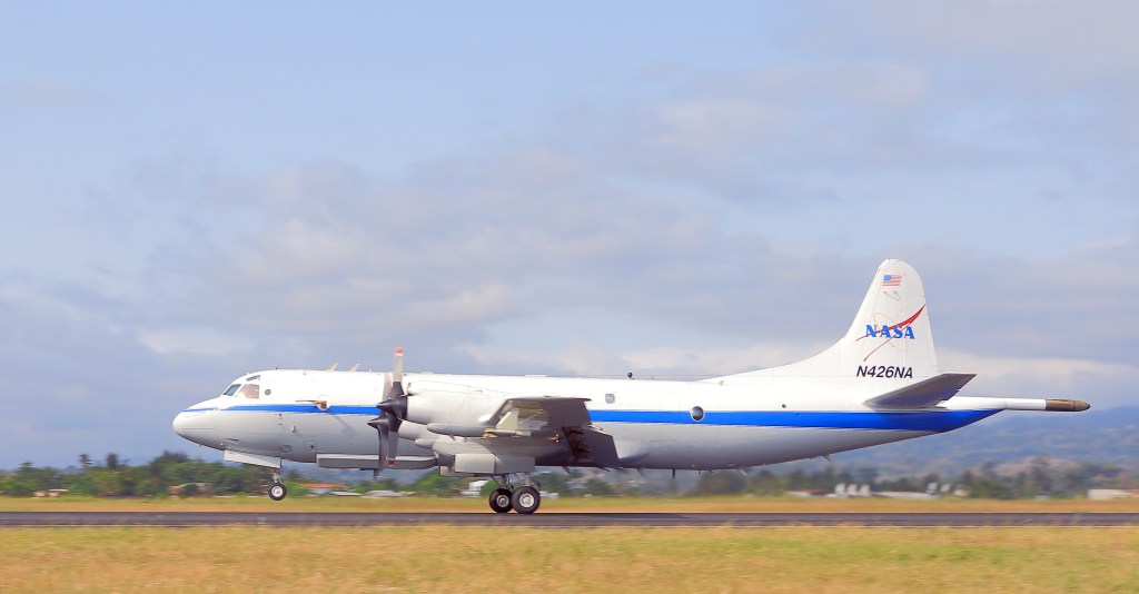 A large propeller plane takes off from a runway, surrounded by brown and green grasses with low buildings out of focus in the background. The plane is white with a blue stripe down the middle, and has a NASA logo on the tail. The sky is a hazy blue with some clouds.