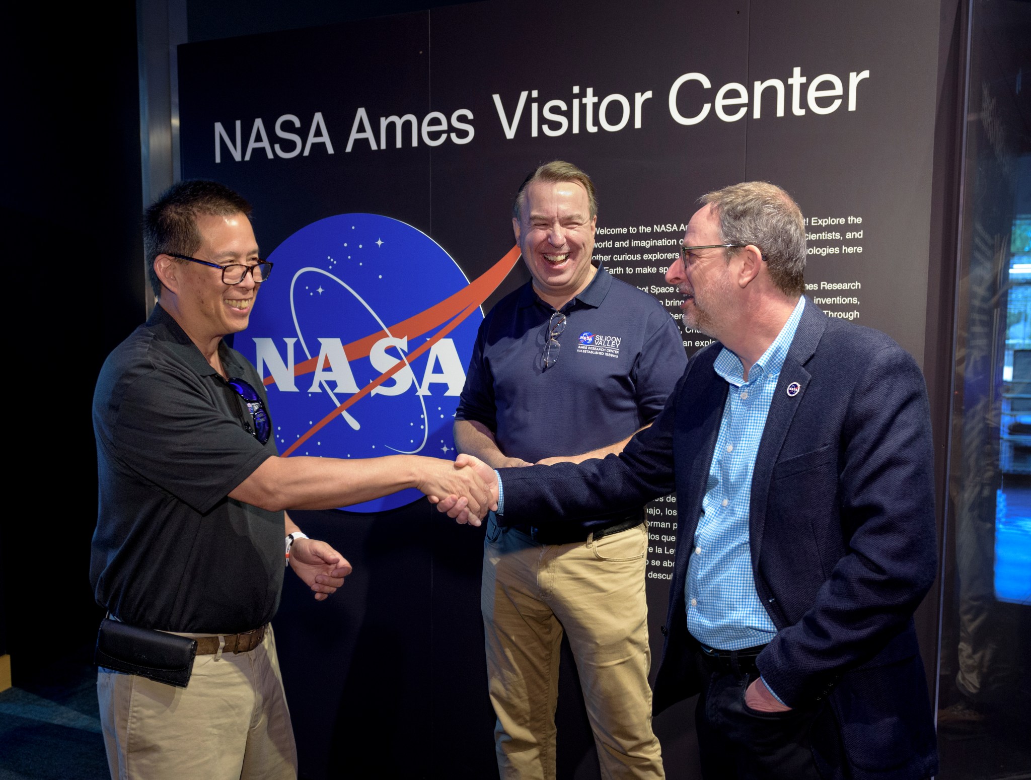 NASA Ames Center Director Eugene Tu, left, shakes hands with Chabot Space & Science Center director Adam Tobin, right, while NASA Ames deputy center director David Korsmeyer, center, looks on. The three individuals are standing in front of the entrance to the NASA Ames Visitor Center exhibit with the NASA meatball logo behind them.