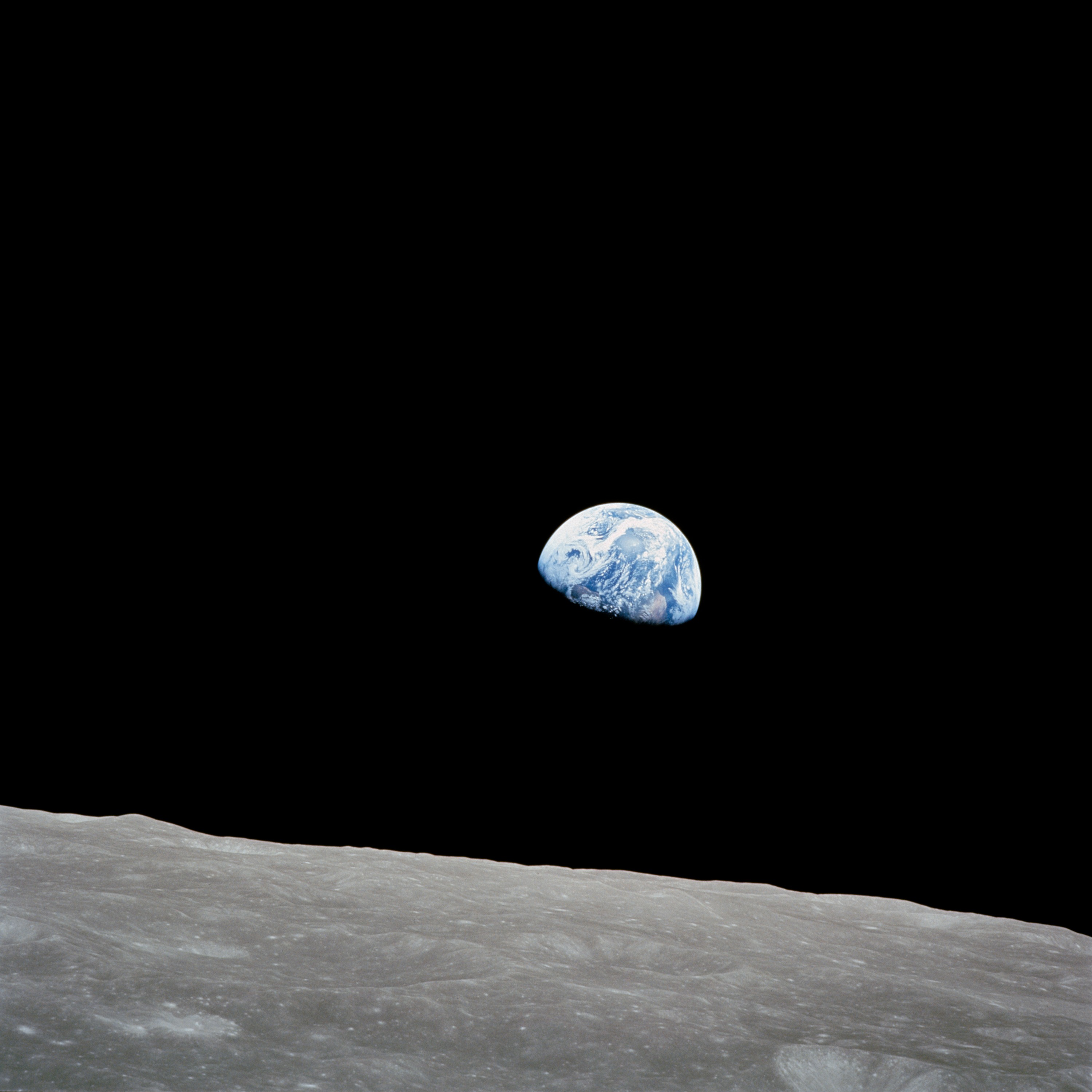 A color photograph of Earth rising over the horizon of the Moon, as seen from the Apollo 8 spacecraft. The Moon's horizon is a dull gray, covering the bottom quarter of the image. Earth, with its blue water and white swirling clouds, is almost at the center of the image. The bottom third of Earth is not visible.