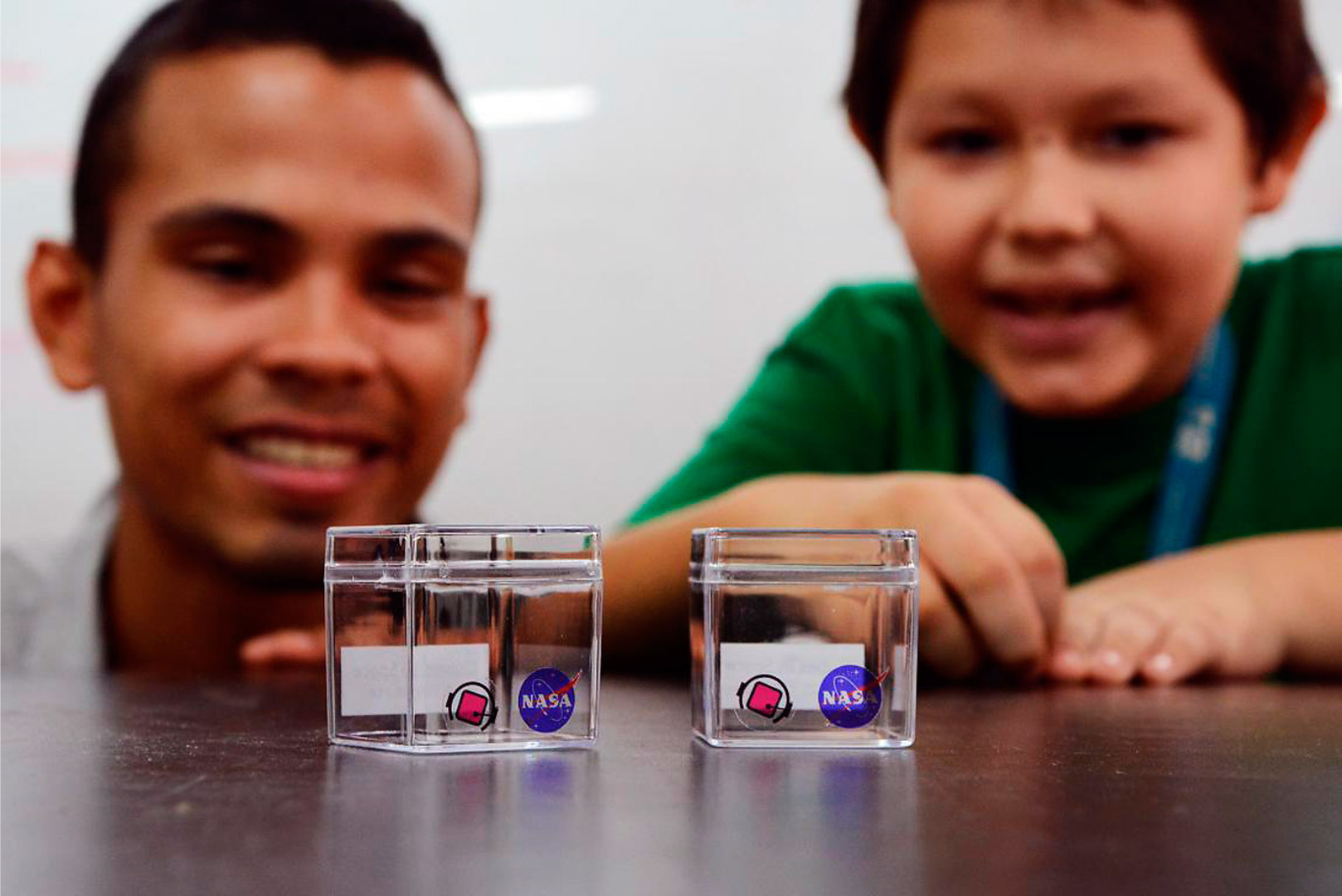 An adult and child are smiling at two clear plastic cube shaped containers that will hold student experiments