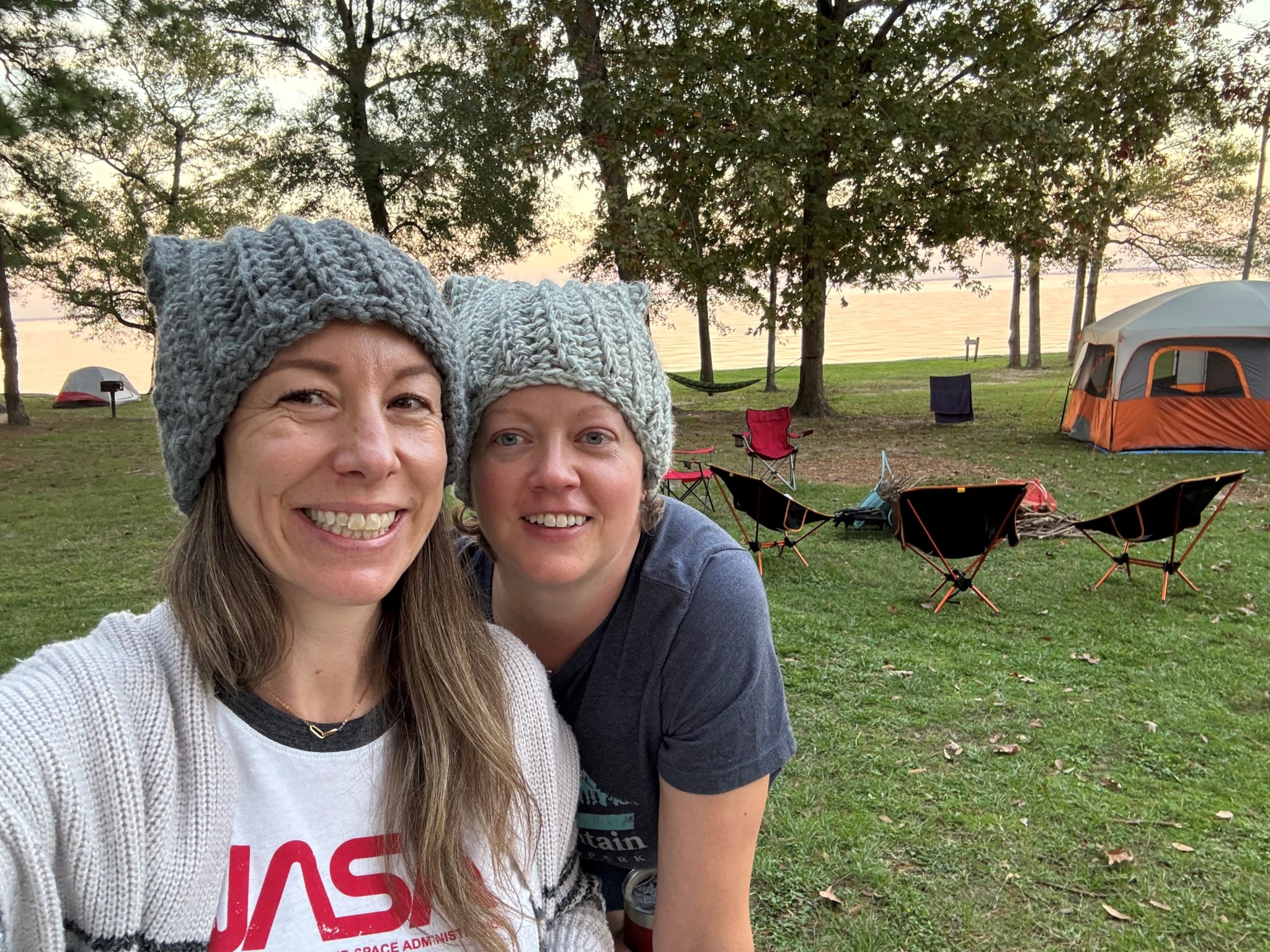 Two women wearing knit hats and t-shirts pose for a photo at a lakeside campsite.
