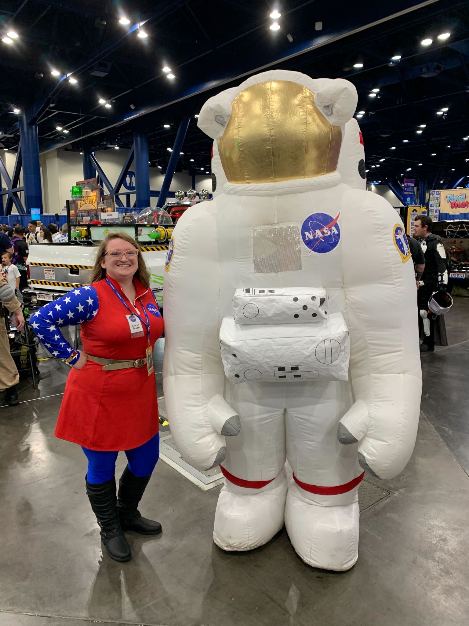 A woman wearing a red, white, and blue dress stands next to an inflatable astronaut mascot at Comicpalooza.