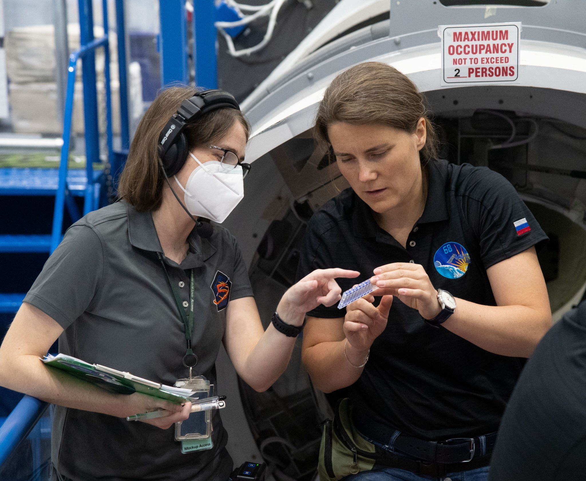 Two people are working together in a facility. The person on the left is wearing a headset, glasses, a face mask, and a gray polo shirt with a logo, and they are holding a clipboard. The person on the right is wearing a black polo shirt with mission patches, including one with a Russian flag, and they are holding a small piece of equipment.
