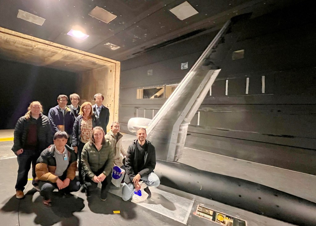 Nine researchers stand in front of a sliver-colored airplane research model in a wind tunnel facility. The model consists of a section of a scaled-down fuselage on the floor, with a single wing mounted vertically on it. The metallic wall of the wind tunnel is visible in the background and a bright light shines in the background.