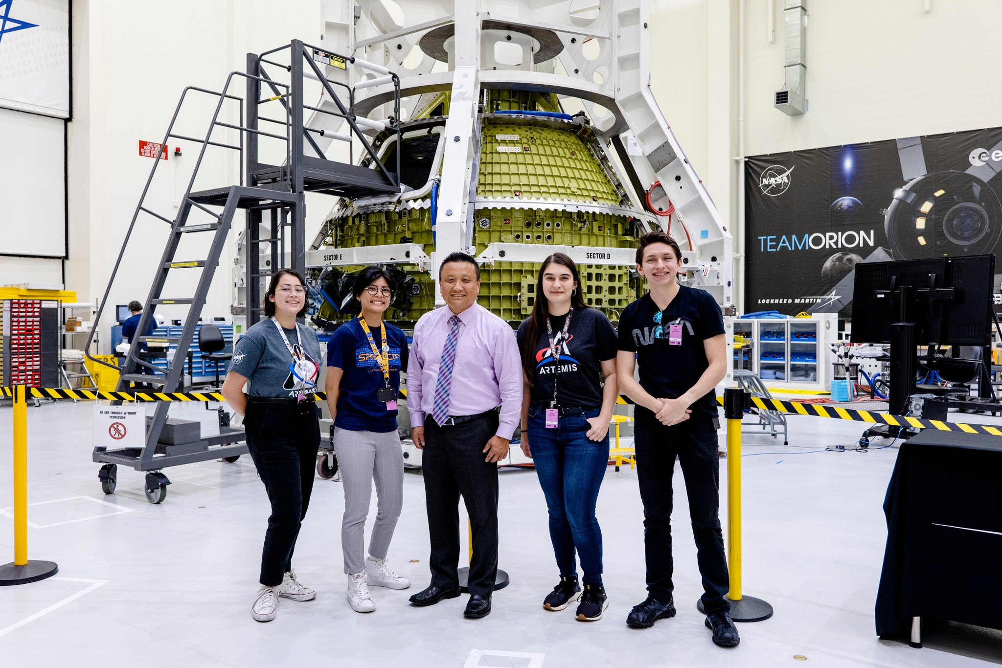 Three diverse youth pose with an Asian American man in business attire in front of the Orion spacecraft crew module.