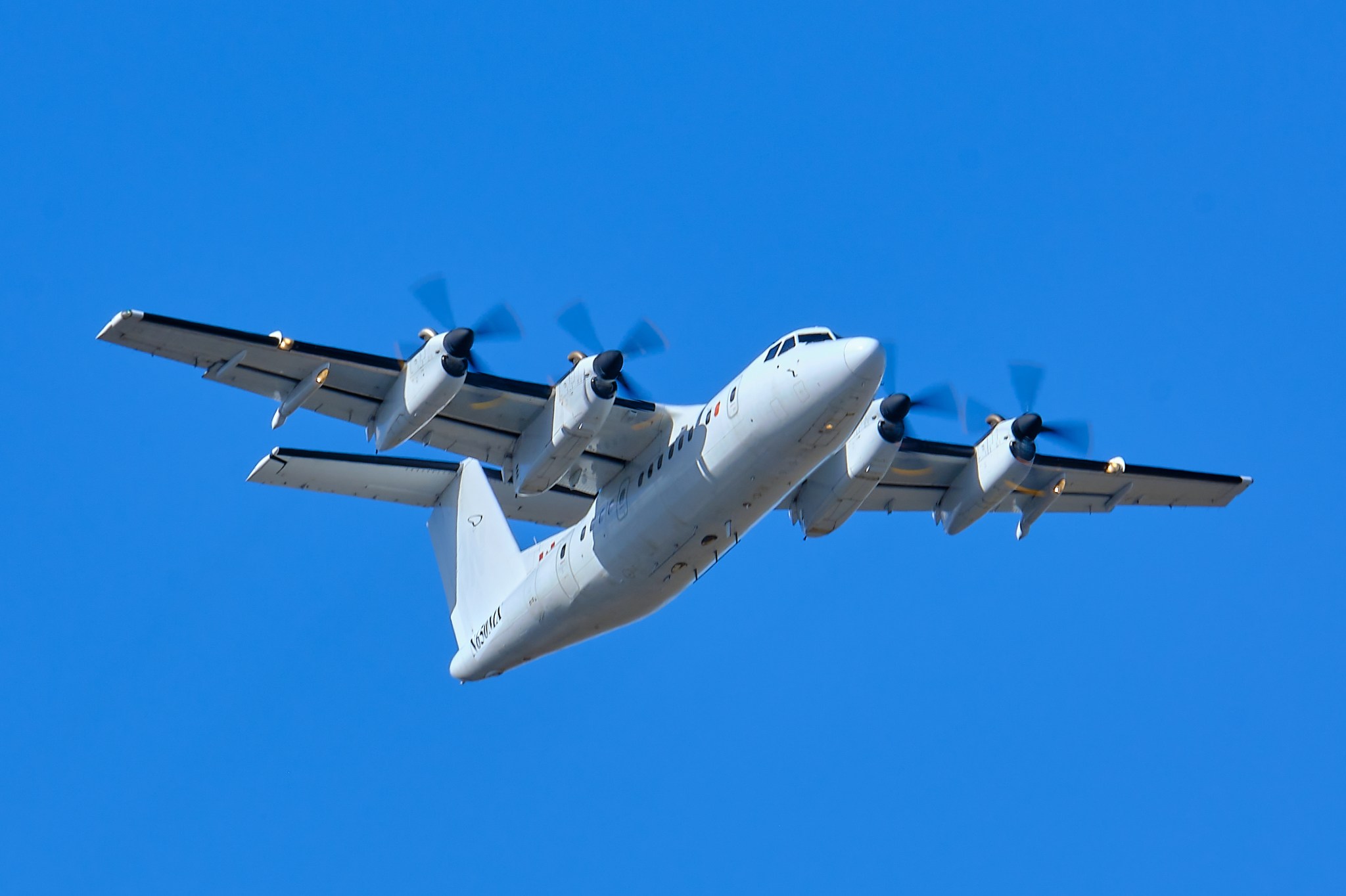 A white turboprop aircraft with two engines under each wing flies across a clear blue sky. The aircraft undergoes baseline flight testing prior to being modified with a hybrid electric propulsion system. 