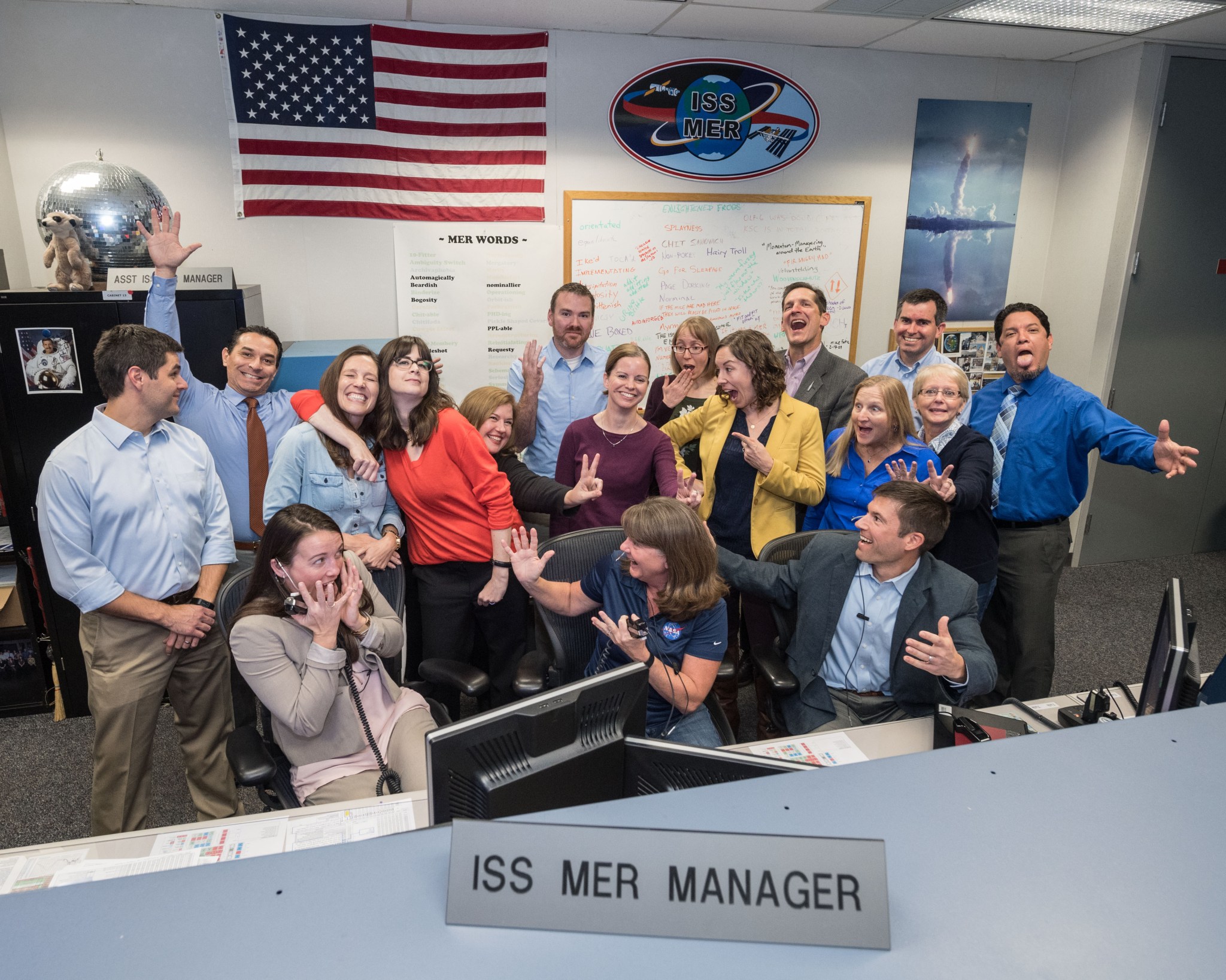 A group of 17 men and women strike silly poses while sitting behind a bank of computers labeled ISS MER Manager.