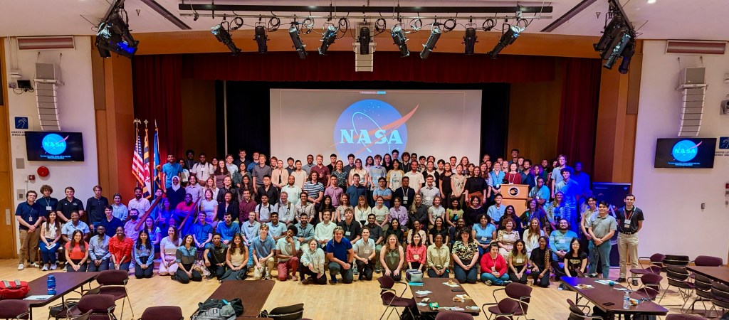 200 interns pose on stage and on the floor in an auditorium. There is a NASA meatball logo projected behind them and on two TVs, one on each side of the stage.