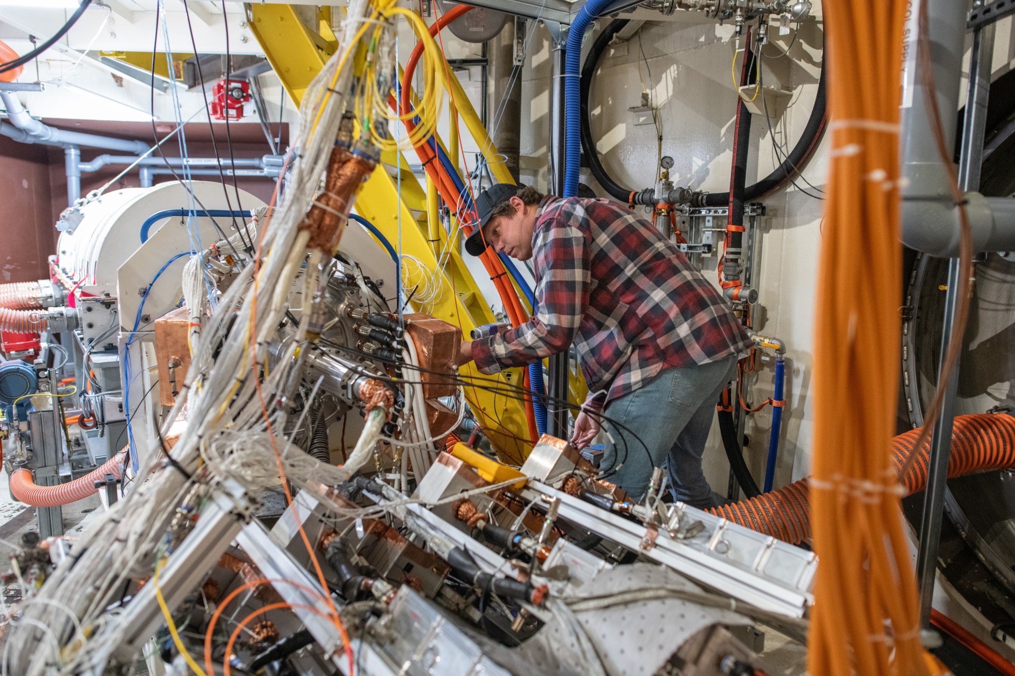 A man in a flannel shirt and jeans accesses a control panel in a room full of wires, cables, and other equipment.