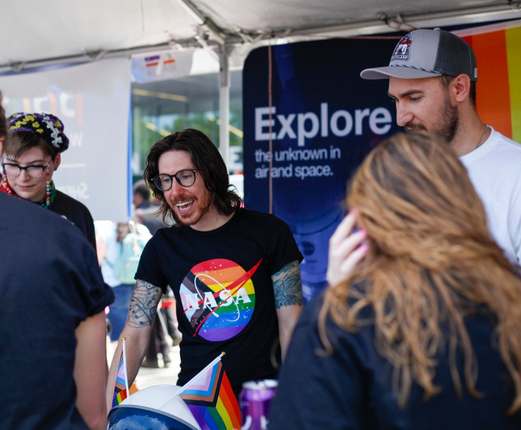 Three NASA Glenn Research Center employees talk with visitors at a table featuring rainbow flags and a NASA space helmet. A colorful banner reading, “Explore the unknown in air and space” hangs in the background.