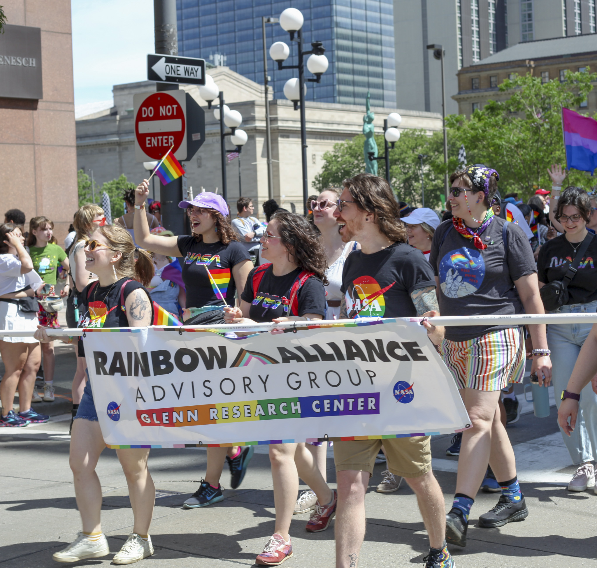 NASA’s Glenn Research Center employees enthusiastically walk in the “Pride in the CLE” parade while wearing colorful shirts and carrying the NASA Glenn Research Center Rainbow Alliance Advisory Group flag in front.