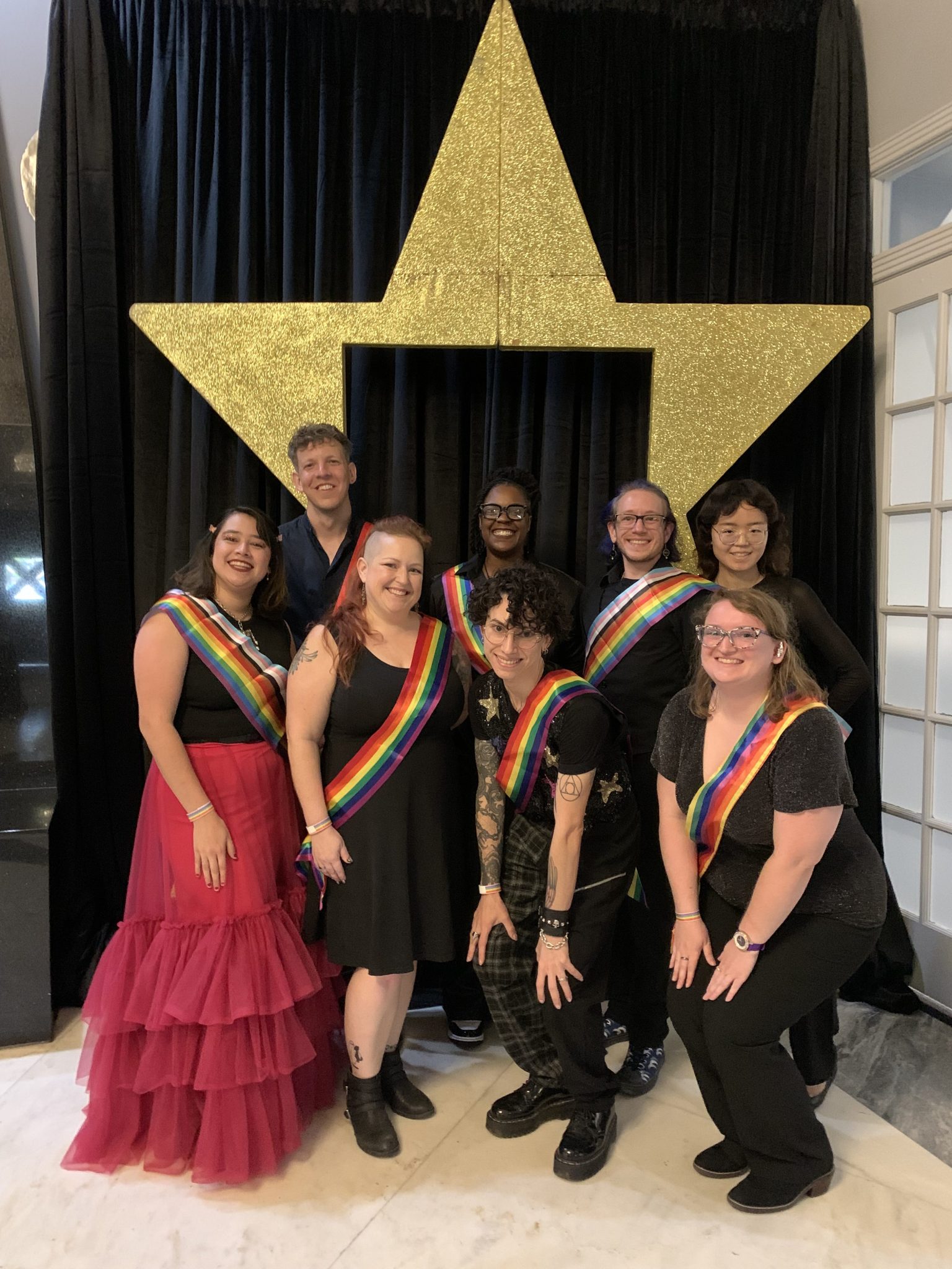 A diverse group of eight individuals wearing rainbow pride sashes pose for a photo in front of a golden star backdrop at a youth prom event.