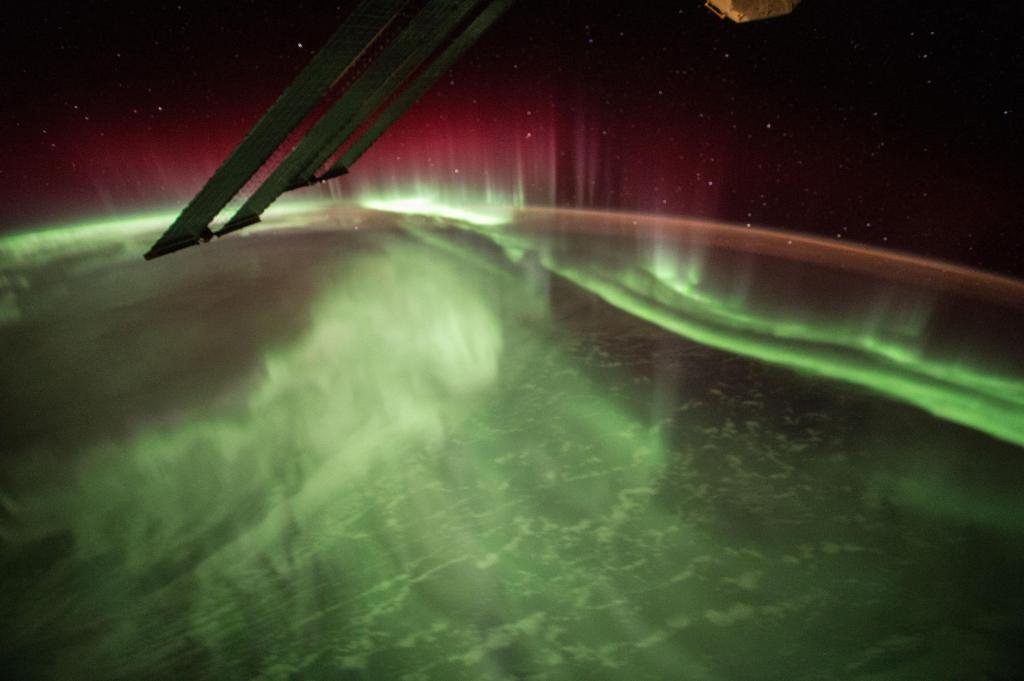Earth’s horizon curves across this image, with a band of reddish light above it and waves of green light across the planet visible below the horizon. Two distinct green streaks of light run from the right side of the image to the middle of the horizon. One of the space station’s solar panels extends down from the top of the image.