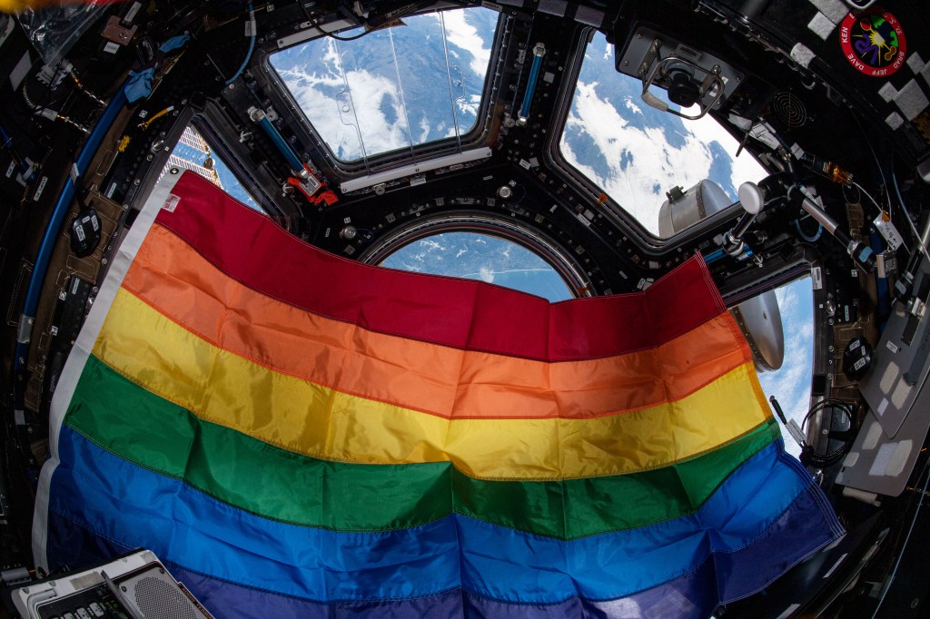 A rainbow flag is displayed inside the cupola aboard the International Space Station, with Earth visible through the windows in the background.