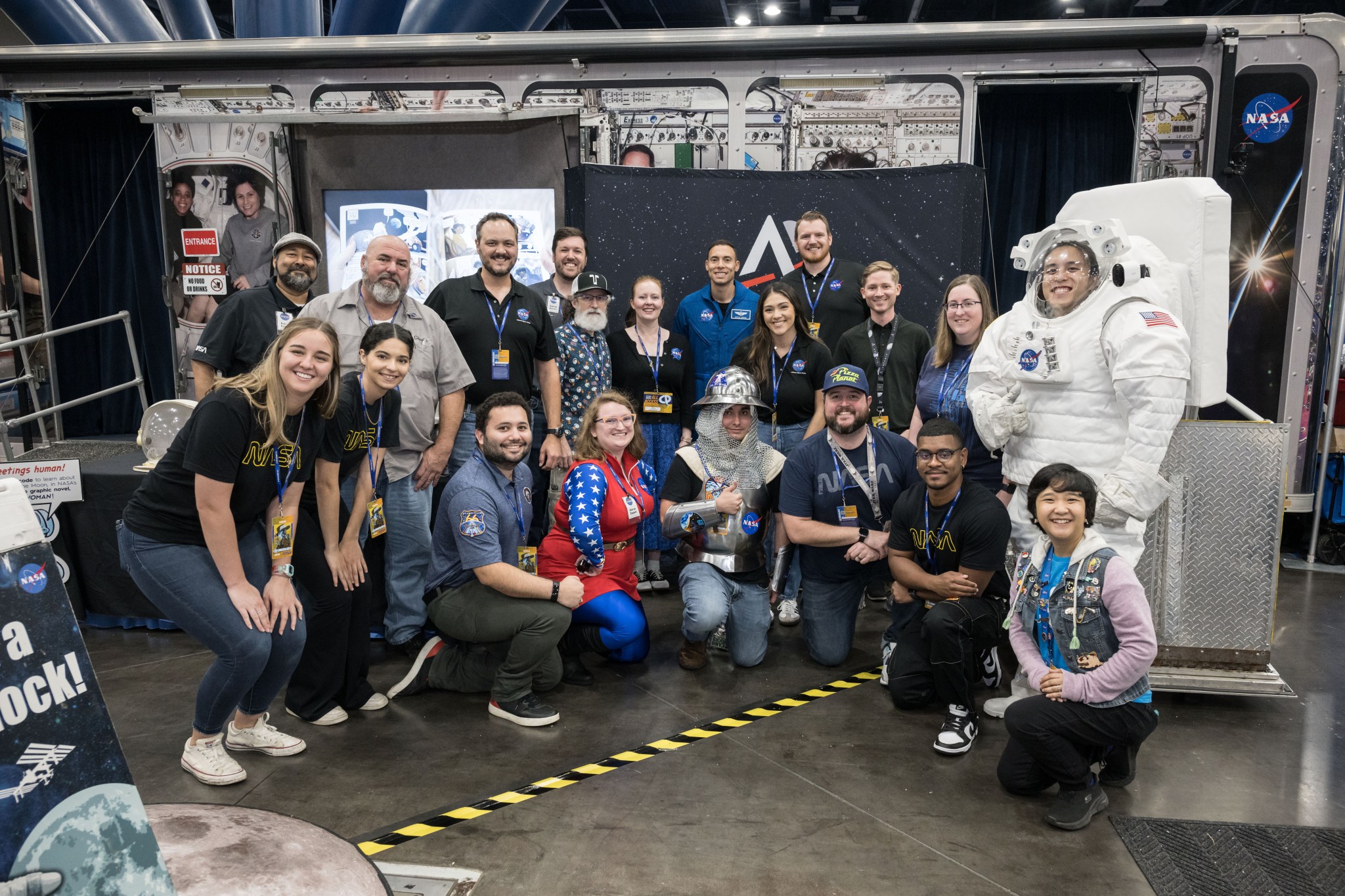 A group of diverse men and women pose for a group photo in front of NASA's exhibit booth at Comicpalooza.