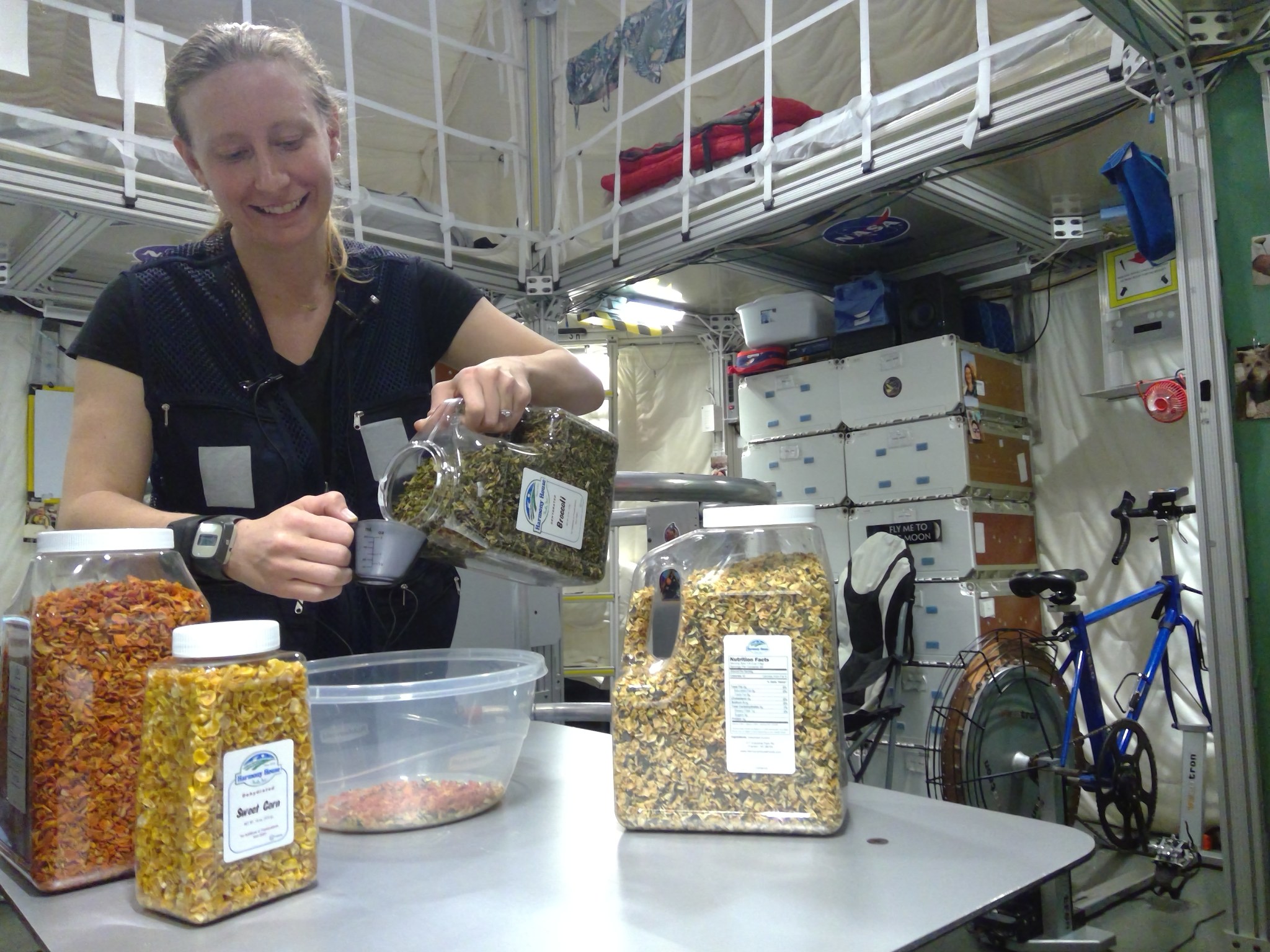A woman in a black uniform pours dried vegetables from a large container into a measuring cup at a table. The table has multiple jars of dried vegetables, and the background features a habitat with storage boxes, a blue exercise bike, and various equipment.