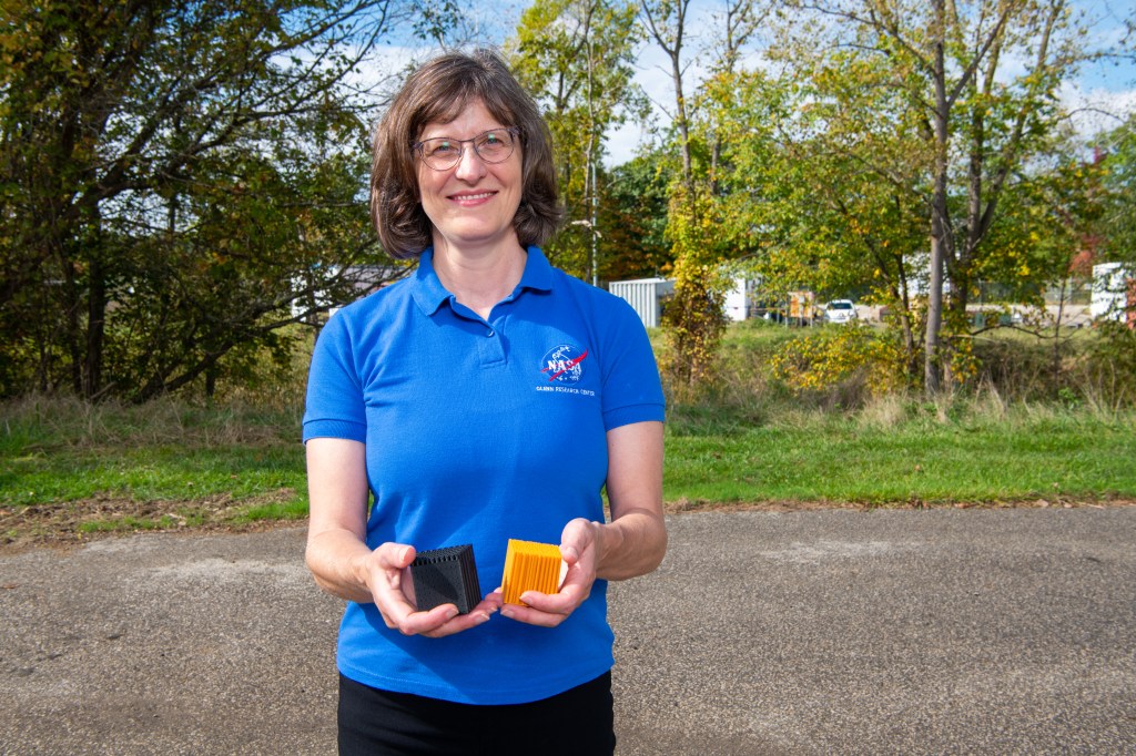 A woman wearing glasses and a blue NASA polo holds up two small cubes, one black and one yellow.