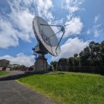 Large dish antenna pointing over trees and up to a blue sky with clouds. Access road and grass in the foreground.