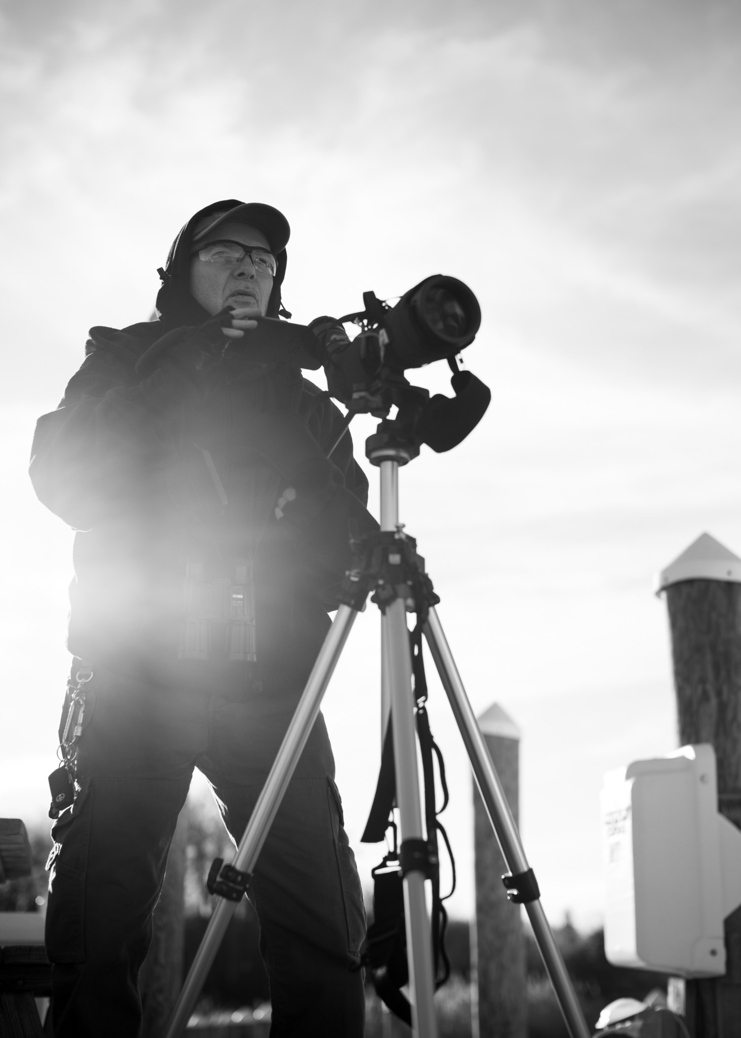 Black and white image of a man operating a camera on a tripod wearing safety glasses, a jacket, pants, and a hat. 