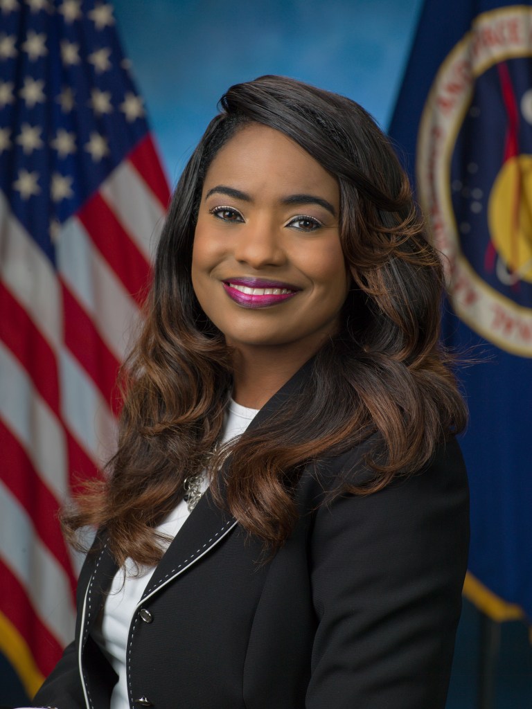 A woman with long, wavy hair and a bright smile poses in front of a U.S. flag and a NASA flag. She is wearing a black blazer with white trim over a white blouse. The background is a gradient blue.