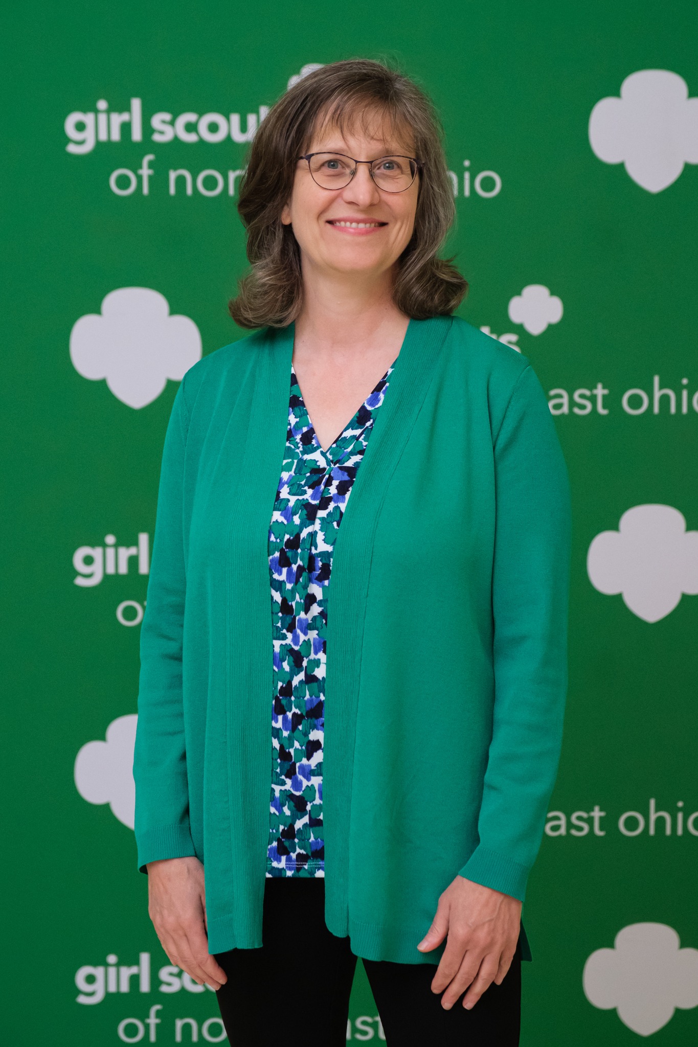 A woman wearing glasses and a blue patterned shirt and green sweater smiles at the camera as she stands in front of a green banner that says, “Girl Scouts of North East Ohio.”