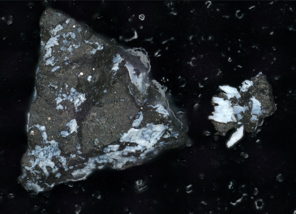 Mineral fragment from OSIRIS-REx's asteroid Bennu sample, seen against a black background. The pieces are predominantly gray, with notable light blue hues flecked throughout. The biggest fragment, triangular, is about a millimeter on a side.