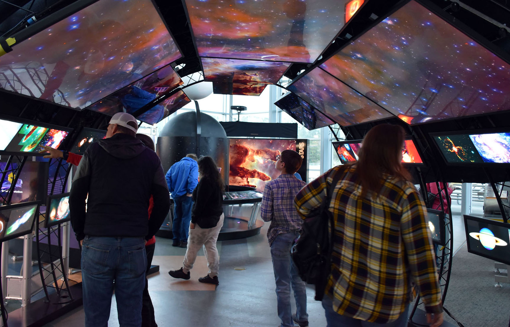 Visitors walk through the Hubble Space Telescope traveling exhibit. An arch of images from the Hubble Space Telescope stretches across the image, from the lower-left, across the top, to the lower-right. People are standing under the arch admiring the images.