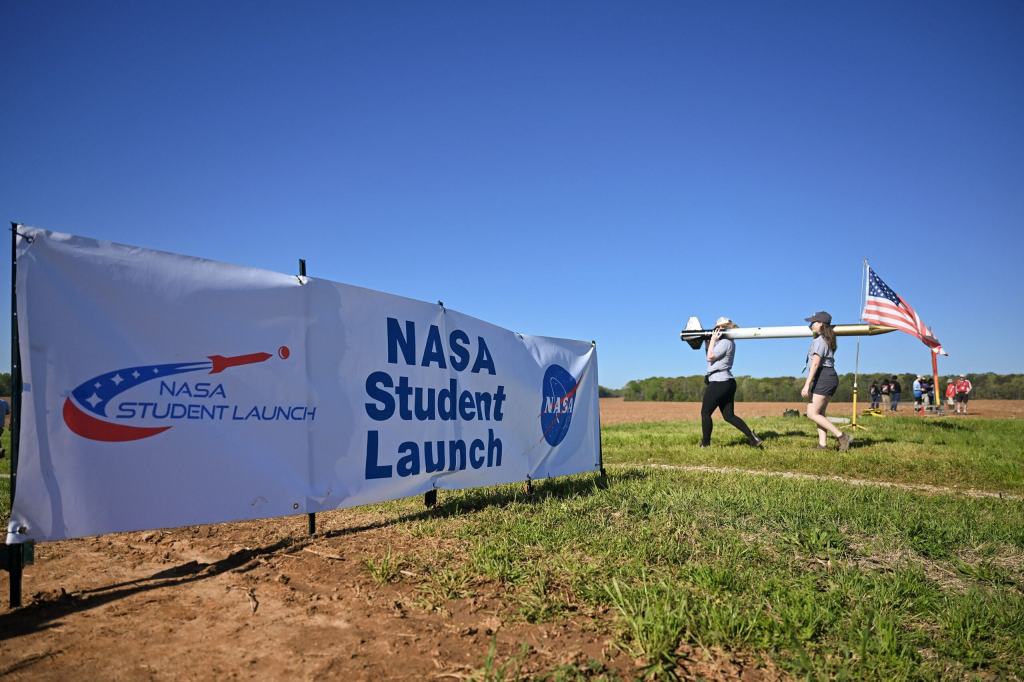 A sign with NASA's Student Launch is in the foreground with two students carrying a rocket in a grassy field.
