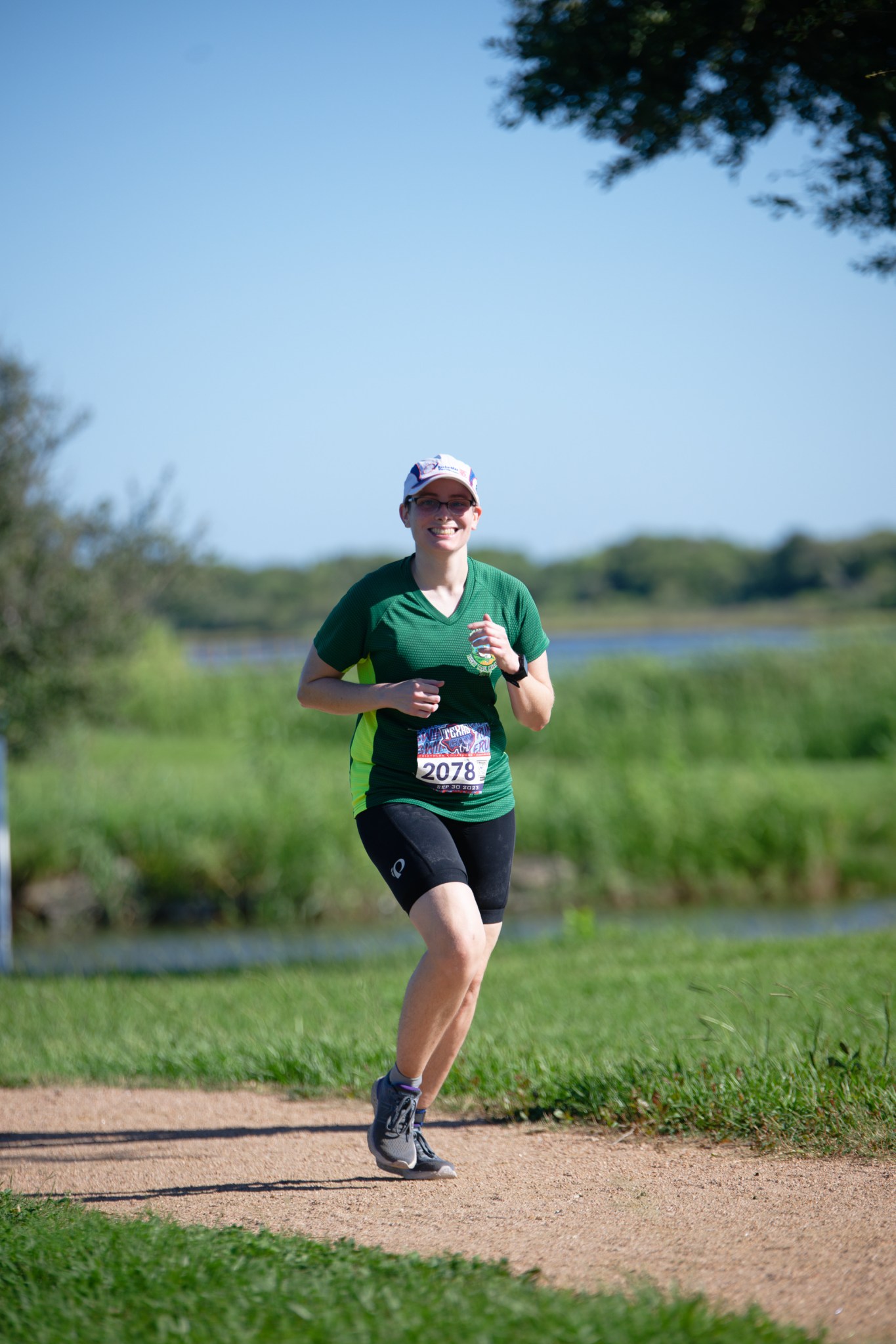 A person is running outdoors on a sunny day. They are wearing a green and black athletic outfit, consisting of a green shirt with short sleeves and black shorts. They have a race bib number 2078 attached to their shirt and are wearing a white cap and glasses.