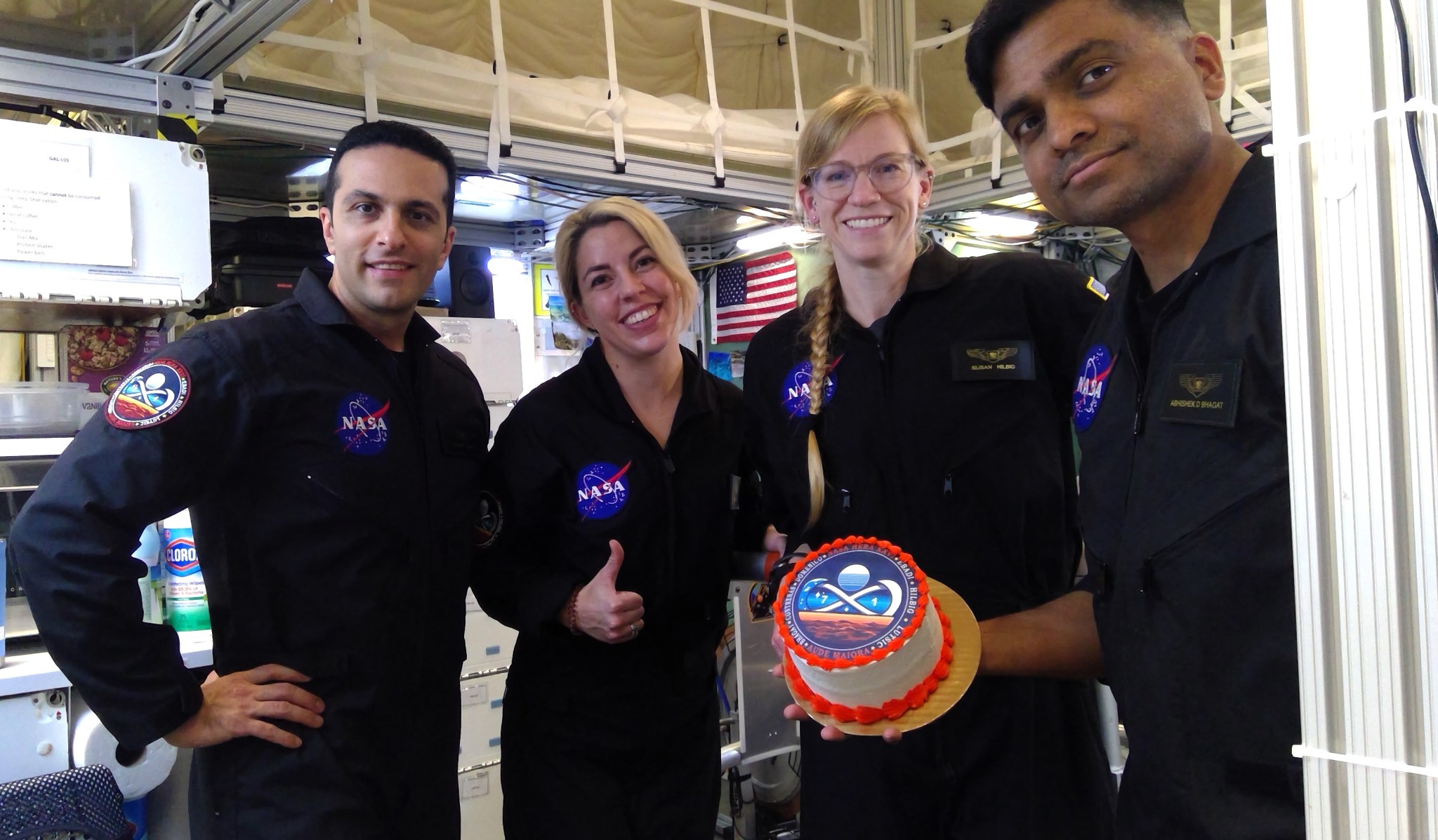 A group of four NASA astronauts in black uniforms are smiling and posing for a photo inside a space station module. One of the astronauts is holding a cake with a NASA logo on it. The background includes various equipment and a U.S. flag.