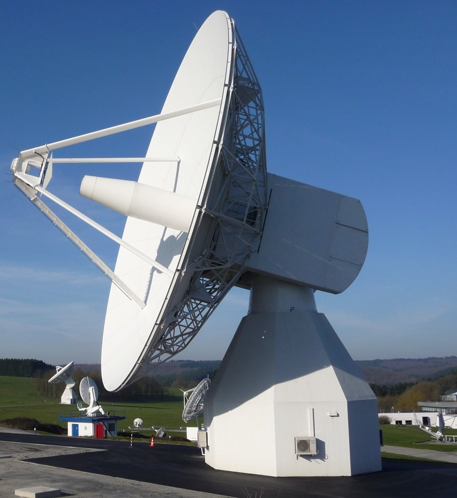 A picture with blue sky and minimal clouds toward the bottom of the image, with a large 20-meter antenna in the forefront, facing the left side of the image.
