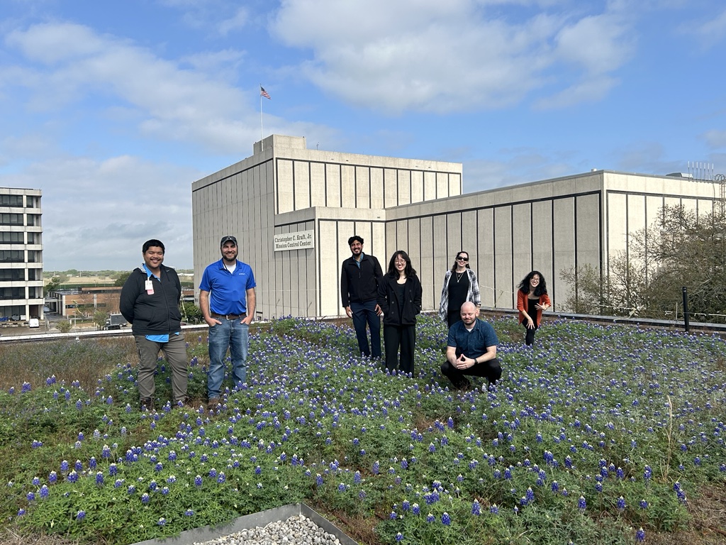 A group of seven people stand and crouch on a rooftop garden filled with blooming bluebonnet flowers. The background includes the Mission Control Center and other buildings, under a partly cloudy sky.