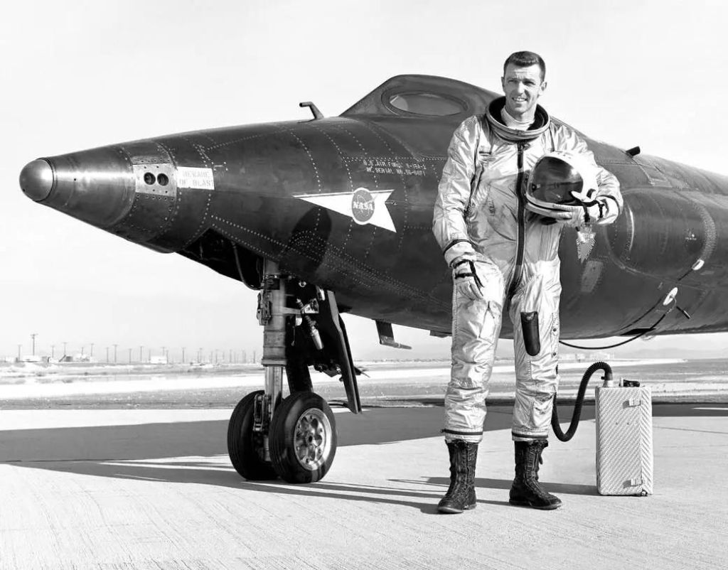 A black-and-white image. Joe Engle stands next to the X-15-2 aircraft. He wears a spacesuit and cradles his helmet with his left arm. There is a hose behind him that attaches to a rectangular box. The plane has a long conical nose and a small window to see out of.