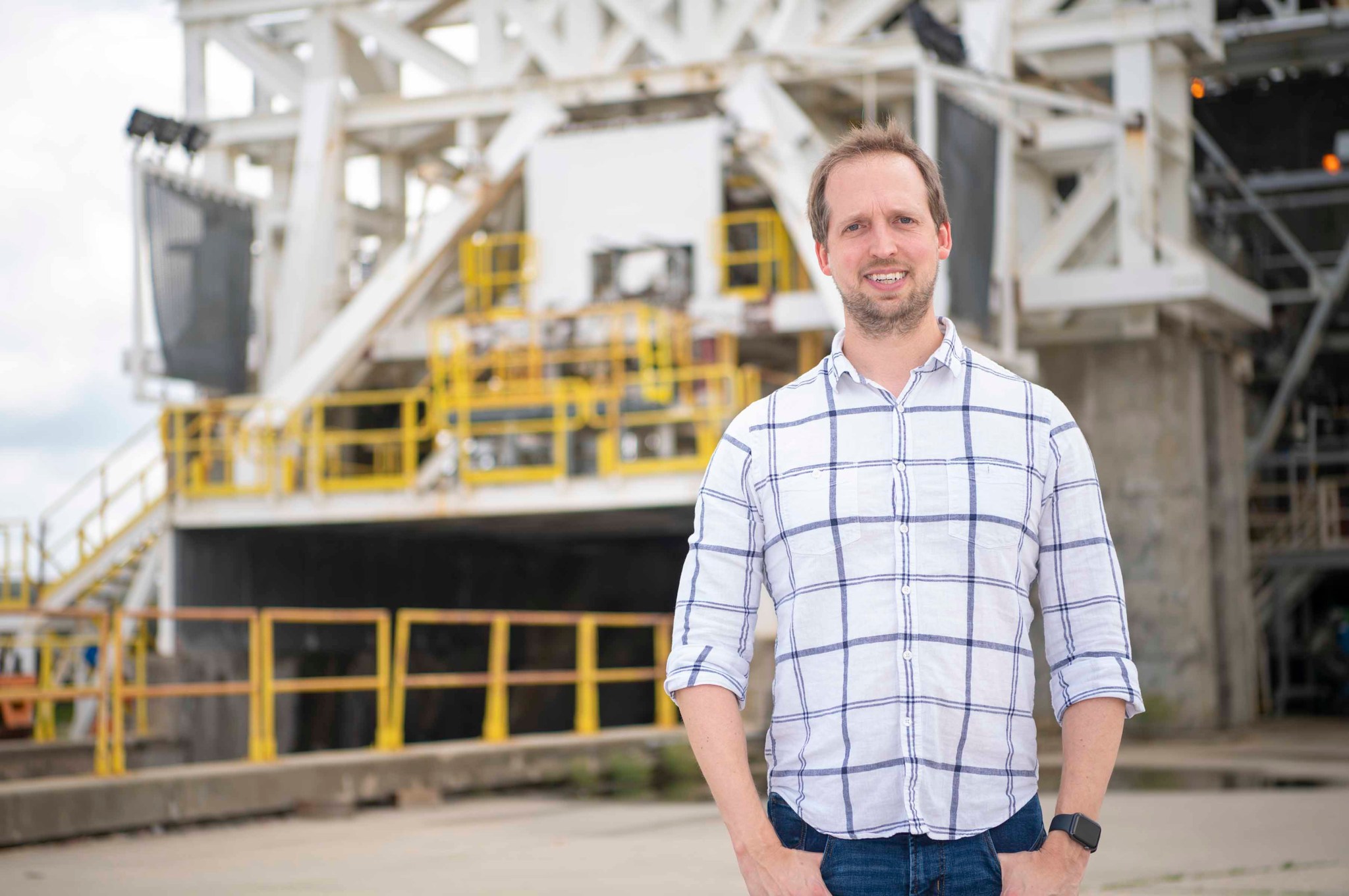 Chris Barnett-Woods, wearing a white dress shirt with black stripes, is shown standing in front of the E-1 Test Stand at NASA’s Stennis Space Center