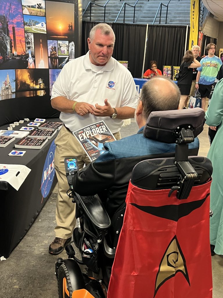 Troy Frisbee, wearing a white polo-style shirt, interacts with a visitor who stopped by the NASA Stennis booth at Mississippi Comic Convention