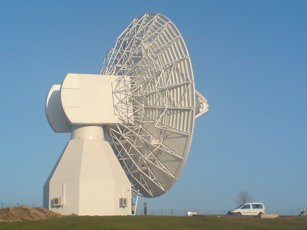 A picture of the backside of a 20-meter antenna designed and built by CPI Satcom in front of a light blue sky. There is a white van toward the right of image — this van looks quite small in comparison to the largeness of the antenna.