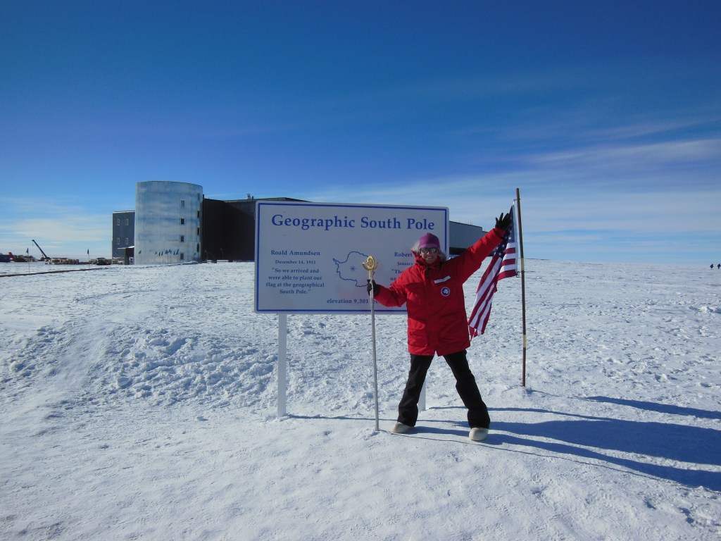 A woman stands smiling at the Geographic South Pole, holding an American flag and a ceremonial staff. She is dressed in a red parka, black pants, white boots, and a pink hat. The background shows a snow-covered landscape with a large building and clear blue sky.