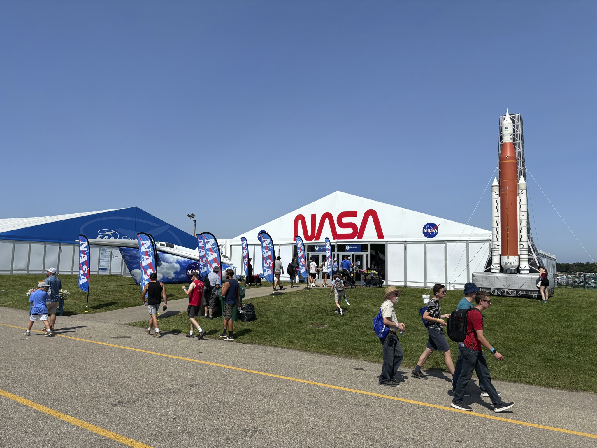 a large white tent with a NASA logo and inflatable Space Launch System rocket and X-59 airplane is in the background as people walk around in the foreground.