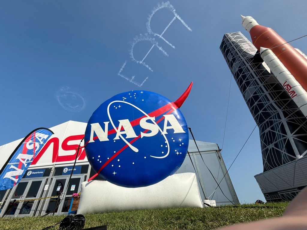 A giant inflatable NASA logo and rocket stand in a front of an exhibit tent beneath clear skies where a skywriting airplane has written EAA and a smiley face.