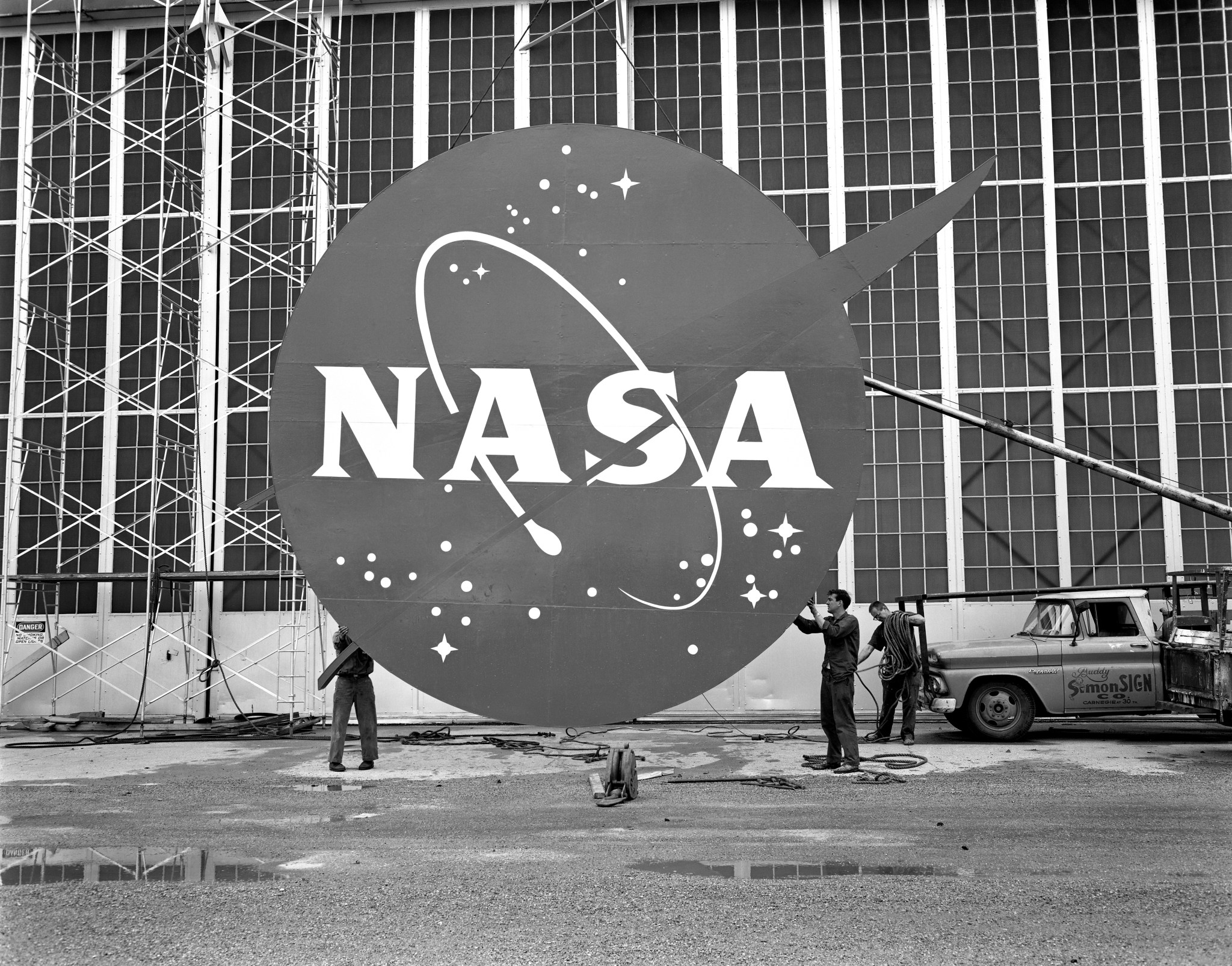In this black-and-white photo, three workers stand below a large NASA logo sign. Two of the workers hold either end of the sign. They stand in front of a hangar building with scaffolding.