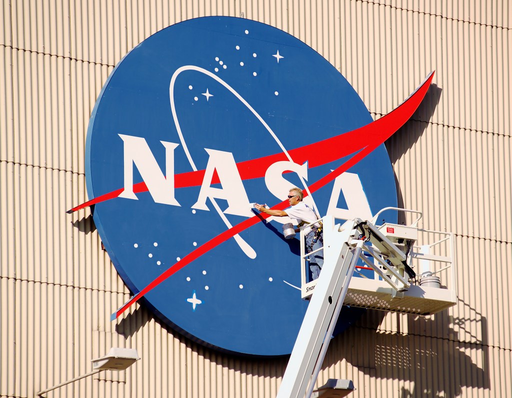 A painter stands in the bucket of a crane lift and reaches out to apply a fresh coat of paint to the red, white, and blue NASA logo painted on a tan, corrugated hangar façade. He is holding a paint brush and a can of paint and wearing a harness, blue jeans, a white shirt, and sunglasses.