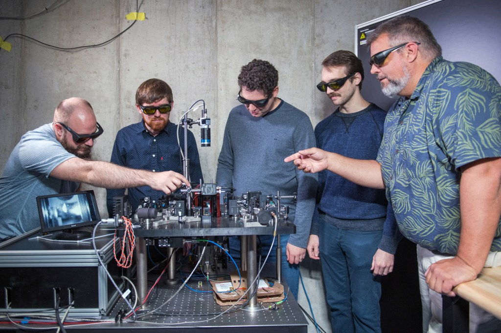 Five engineers inspect laboratory equipment. The quantum memory the equipment holds is shown on a digital screen. Engineers wear eye protection and point at the equipment.