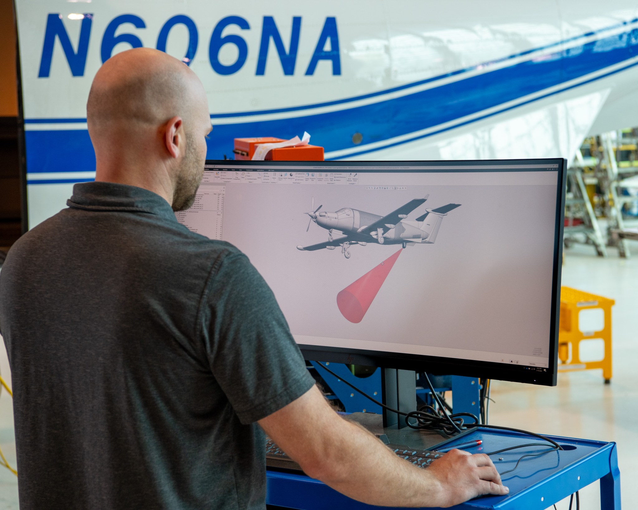 A man wearing a dark grey T-shirt is located on the left side of this image. We see the back of his head, staring at a large computer monitor in front of him. The monitor displays an image of the PC-12 aircraft, with a red laser signal shooting out of the bottom of the aircraft.