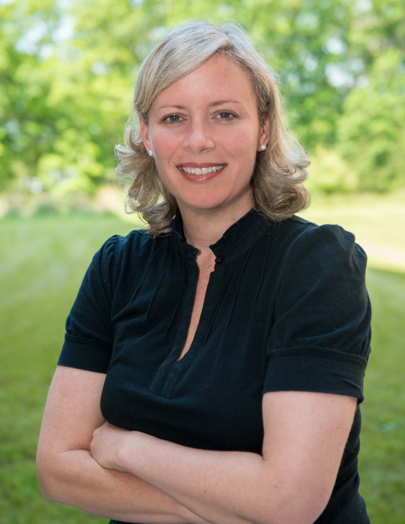 A woman with light blonde hair, wearing a black short-sleeved blouse, stands with her arms crossed outside during the day with trees behind her. She is smiling at the camera.