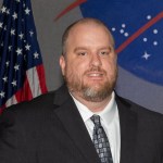 A white man with a graying beard poses in a suit and tie while sitting in front of the America and NASA flags