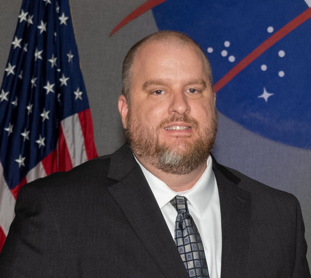 A white man with a graying beard poses in a suit and tie while sitting in front of the America and NASA flags