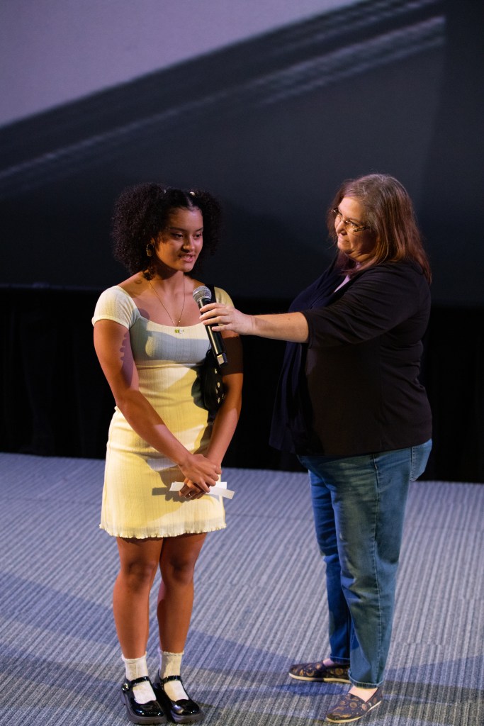 A young woman stands on a stage, holding a piece of paper. An adult woman beside her is holding a microphone. The young woman wears a light-colored dress and black shoes with white socks while the adult woman is dressed in jeans and a black top.