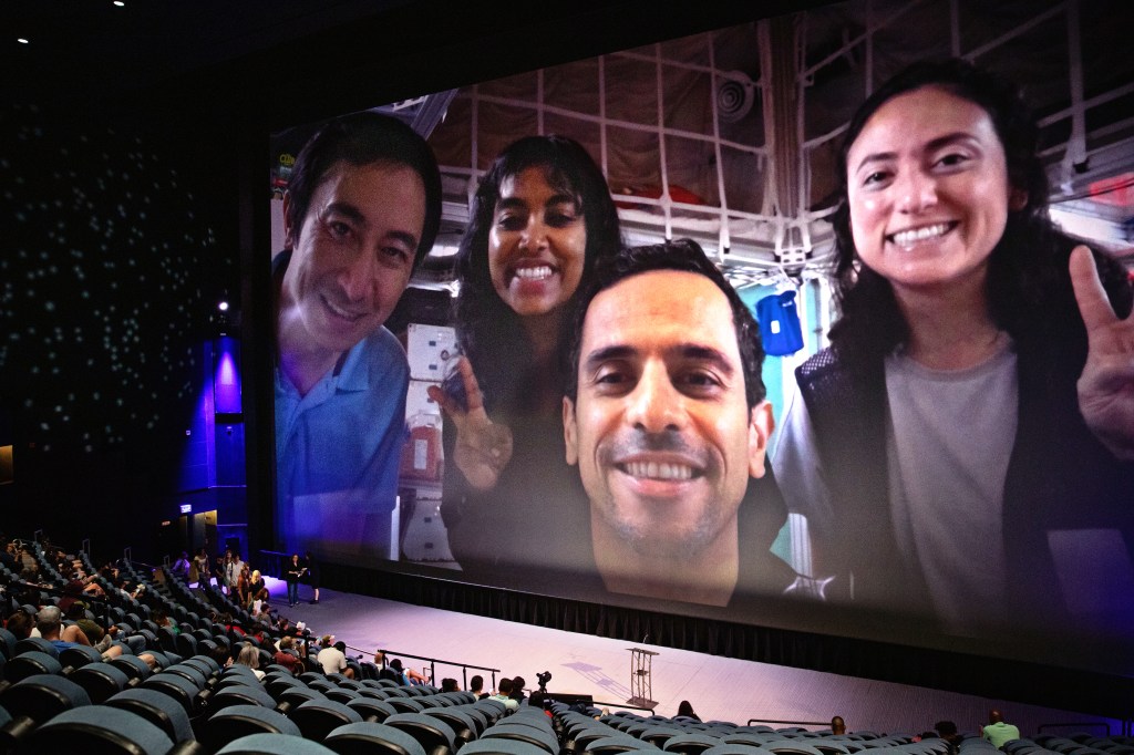 A large screen in an auditorium displays a close-up selfie of four smiling NASA personnel. They are in a spacecraft interior, with equipment visible in the background. One person is making a peace sign. The auditorium seats are partially filled with attendees watching the presentation.