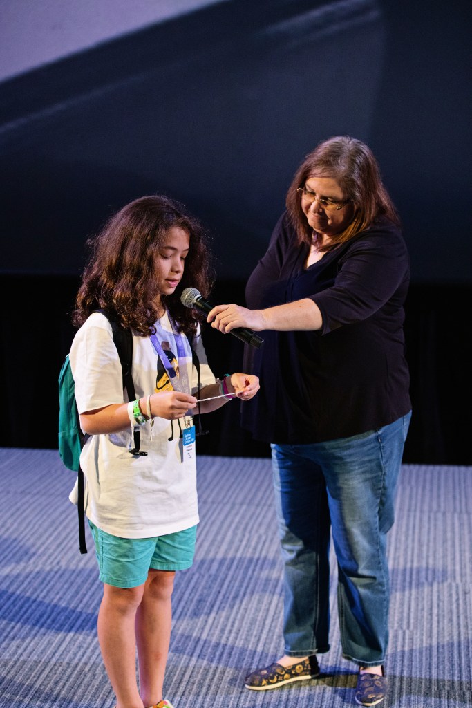 A kid with long hair and a backpack stands on a stage, reading from a piece of paper. An adult woman beside them holding a microphone. The kid wears a white t-shirt and turquoise shorts, while the woman is dressed in jeans and a black top.