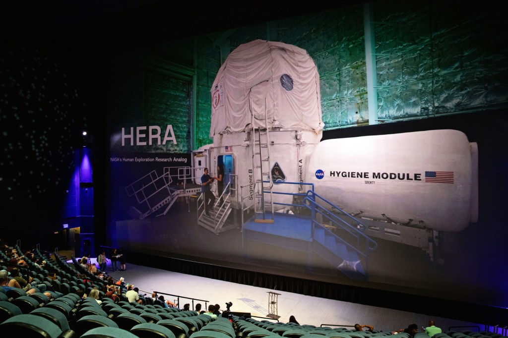 A large screen in an auditorium displays an image of the HERA (Human Exploration Research Analog) habitat module with a "Hygiene Module" labeled next to it. The image shows the white cylindrical habitat with attached modules, metal staircases, and platforms. The background has green lighting and an insulated wall.
